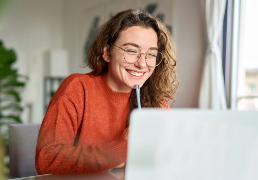 Happy young woman using laptop sitting at desk writing notes while watching webinar, studying online, looking at pc screen learning web classes or having virtual call meeting remote working from home.